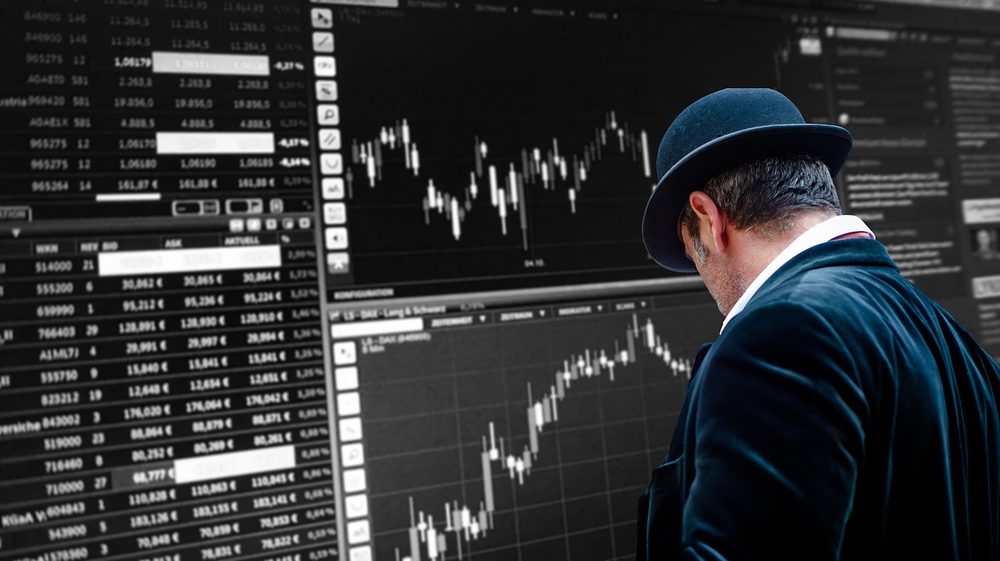 黒スーツの男性が電光掲示板で株価チャートを見つめるイメージ｜イナゴ投資家の定義を示す写真
